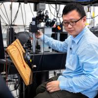 Wei Wang, in a blue shirt and black plastic-framed glasses, sits in a lab looking at an instrument while he adjusts another instrument with his right hand.